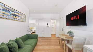 A modern living room with a green sofa, a dining area with a wooden table, and a flat-screen TV mounted on the wall displaying 'NETFLIX'.