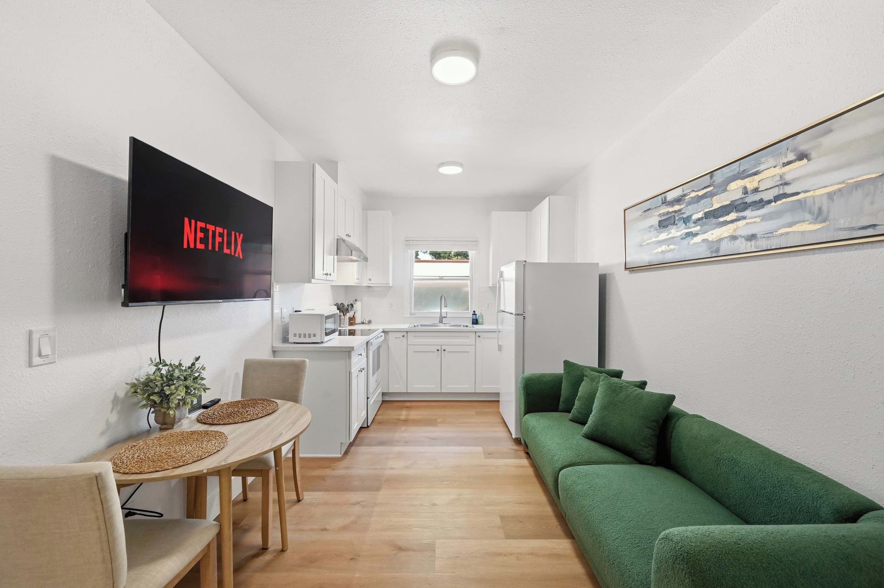 Un espacio habitable compacto con kitchenette, una zona de comedor con mesa y sillas, un sofá verde y un televisor de pantalla plana montado en la pared.