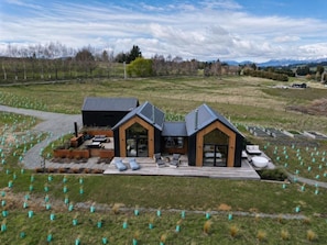 A modern house with a deck, large windows, and a view of a field with trees. - Kākāpō Nest | Sauna, Outdoor Bath, Mountain Views (Te Anau)