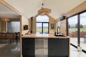 A modern kitchen with a large island, black cabinetry, and a view of a vineyard. - Kākāpō Nest | Sauna, Outdoor Bath, Mountain Views (Te Anau)