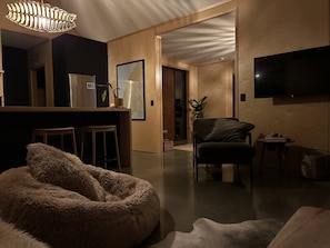 A modern living room with a sofa, a television, and a kitchen area in the background. - Kākāpō Nest | Sauna, Outdoor Bath, Mountain Views (Te Anau)