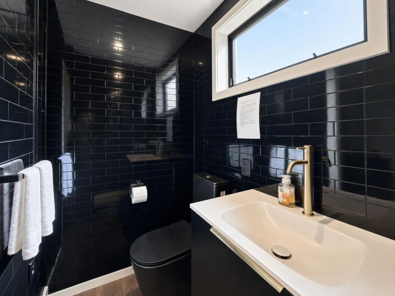 A modern bathroom with black tiles, a white sink, and a window above the mirror.