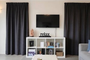 A living room with a flat-screen TV mounted on the wall, a white entertainment unit with books and decorative items, and black curtains on either side. - Beyond the Bay | Rural Riverton Escape (Riverton/Aparima)