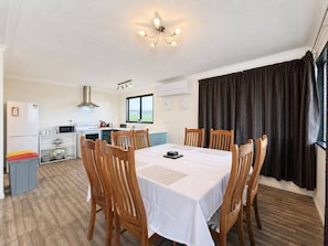 Un coin repas avec une table dressée pour six personnes, entourée de chaises en bois, une cuisine en arrière-plan et une grande fenêtre avec des rideaux.