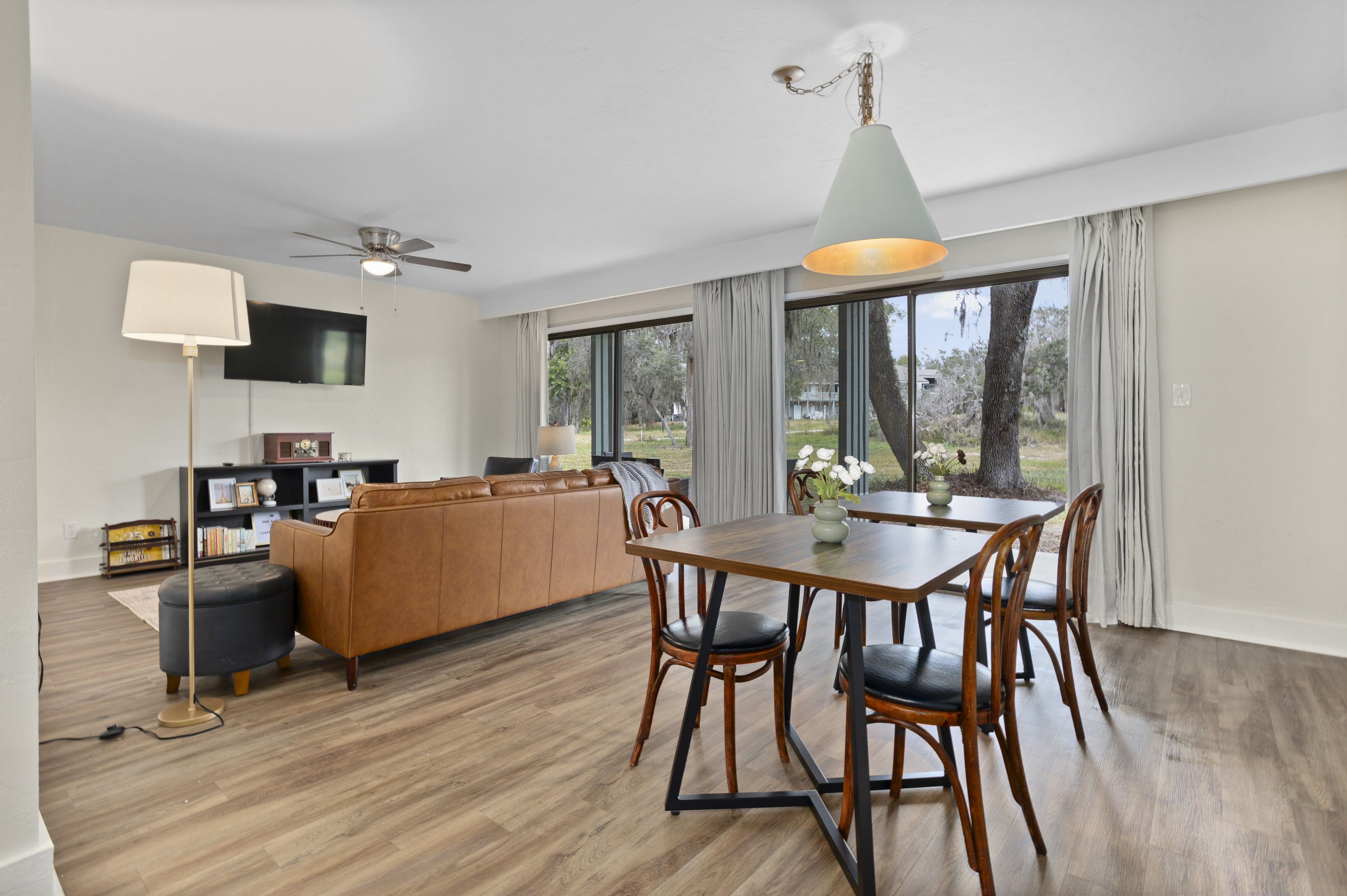 A modern living room with a sofa, a dining table with chairs, a ceiling fan, and a television mounted on the wall.