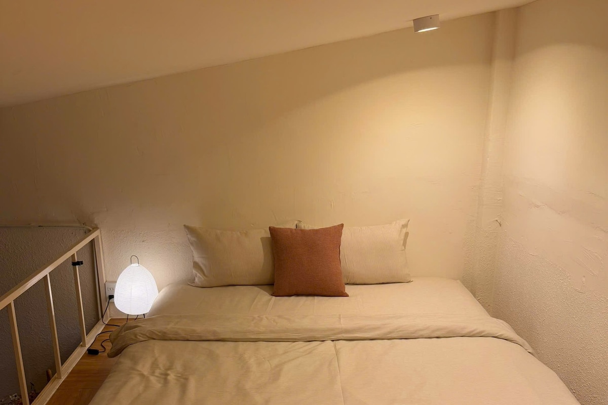 Pequeña habitación con cama individual, suelo de madera, mesita de noche con lámpara e iluminación en la pared.