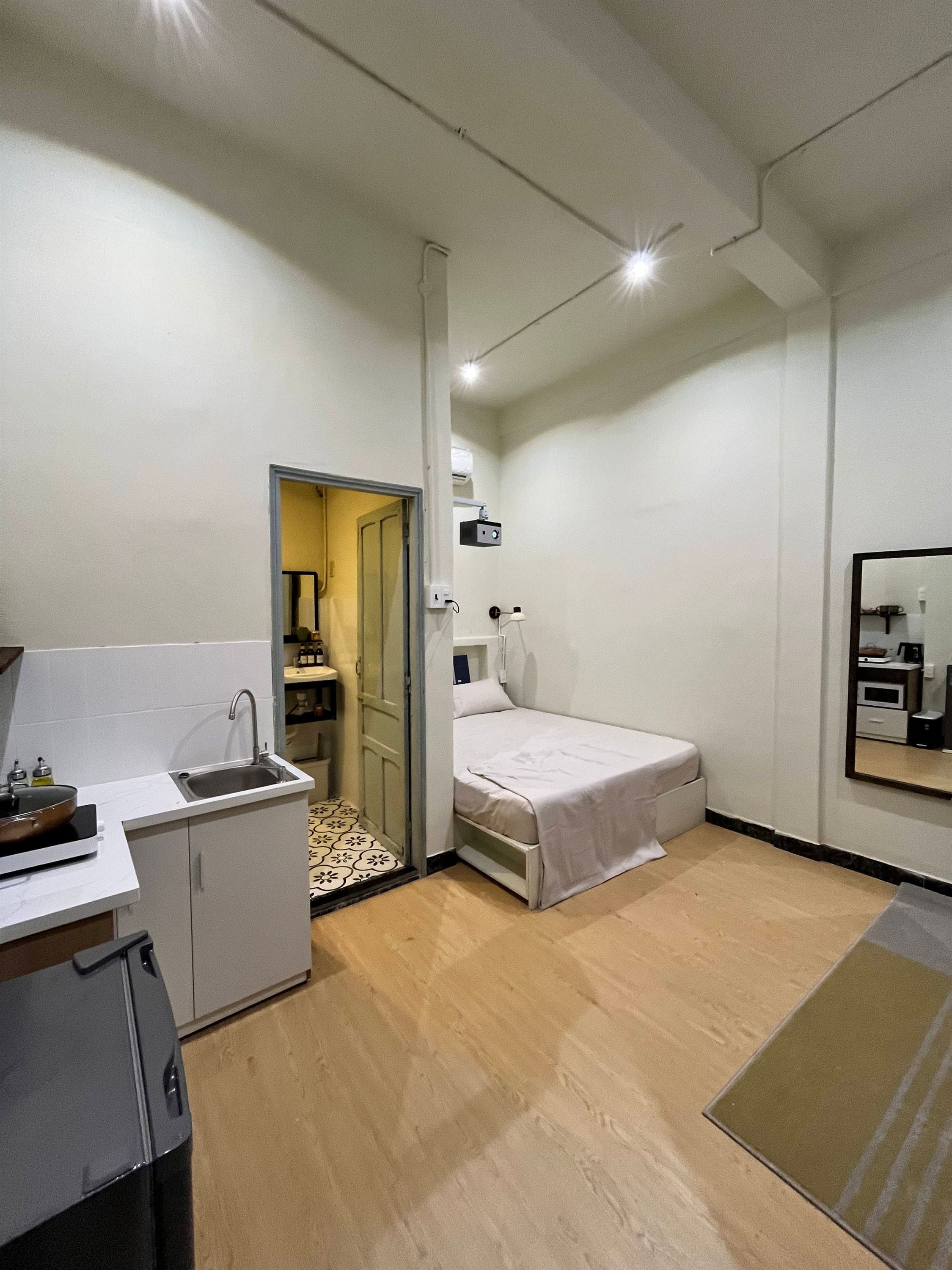 Habitación de hotel pequeña con kitchenette, una cama individual, baño con espejo y puerta que da a otra habitación.