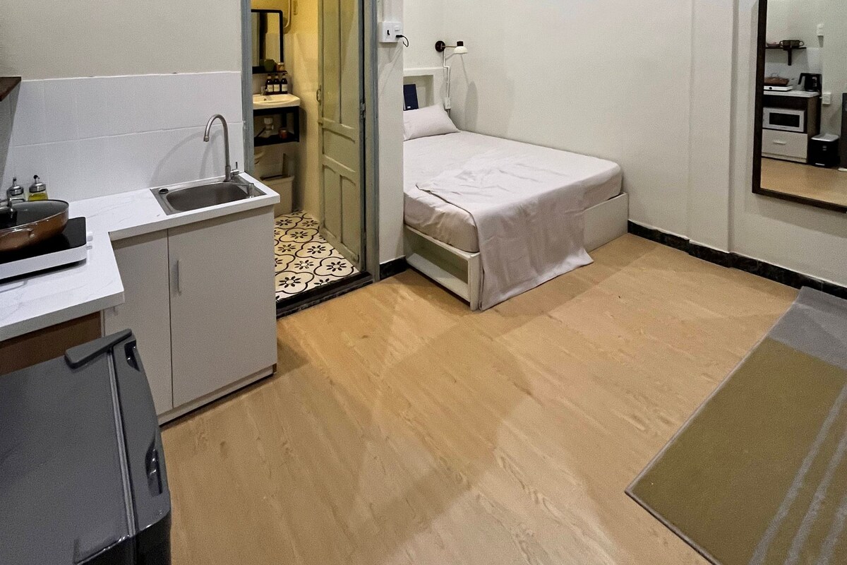 Habitación de hotel pequeña con kitchenette, una cama individual, baño con espejo y puerta que da a otra habitación.