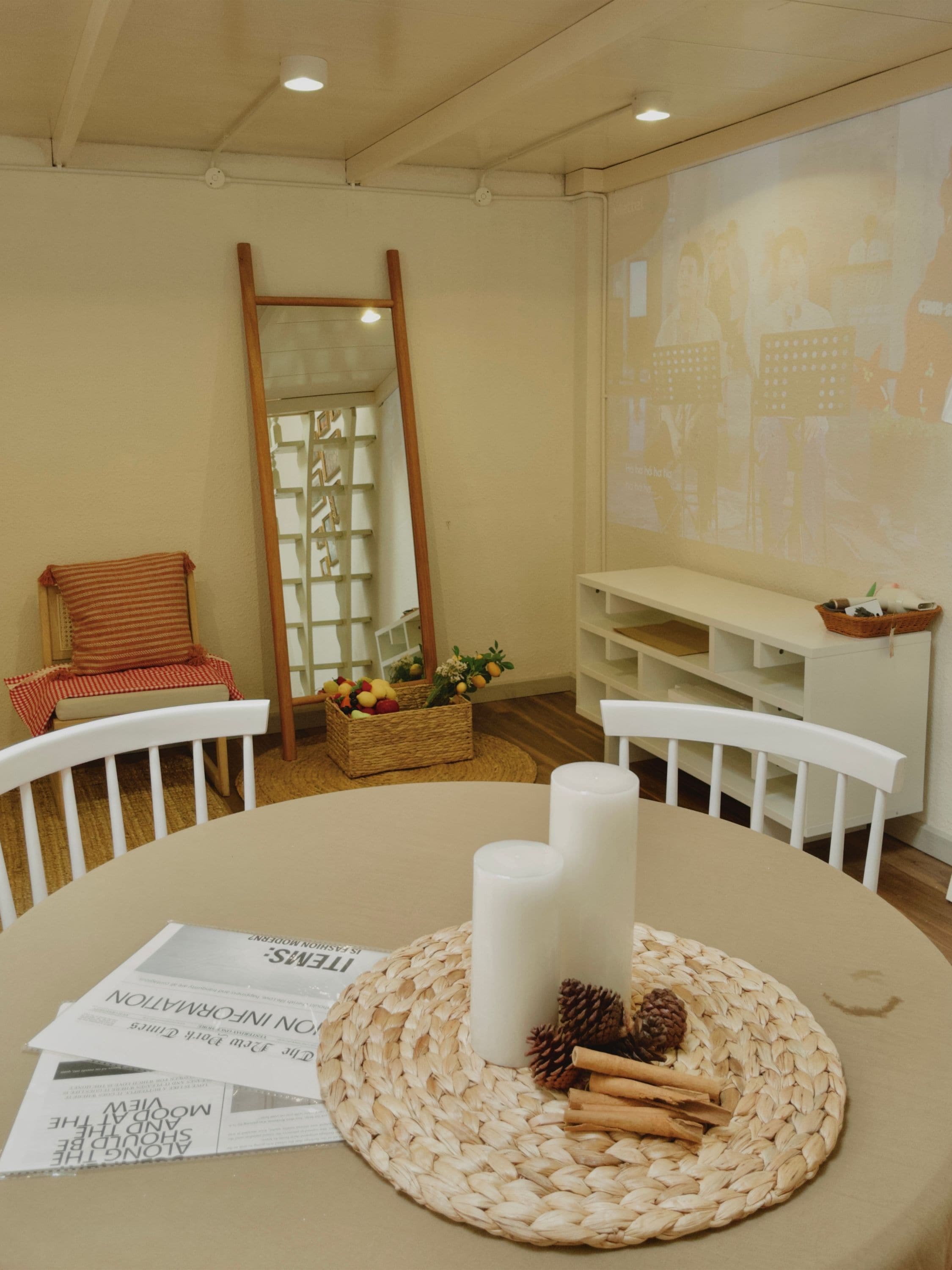 Una habitación con una mesa redonda, sillas blancas, un mantel tejido, velas y una pantalla de proyector mostrando una presentación.