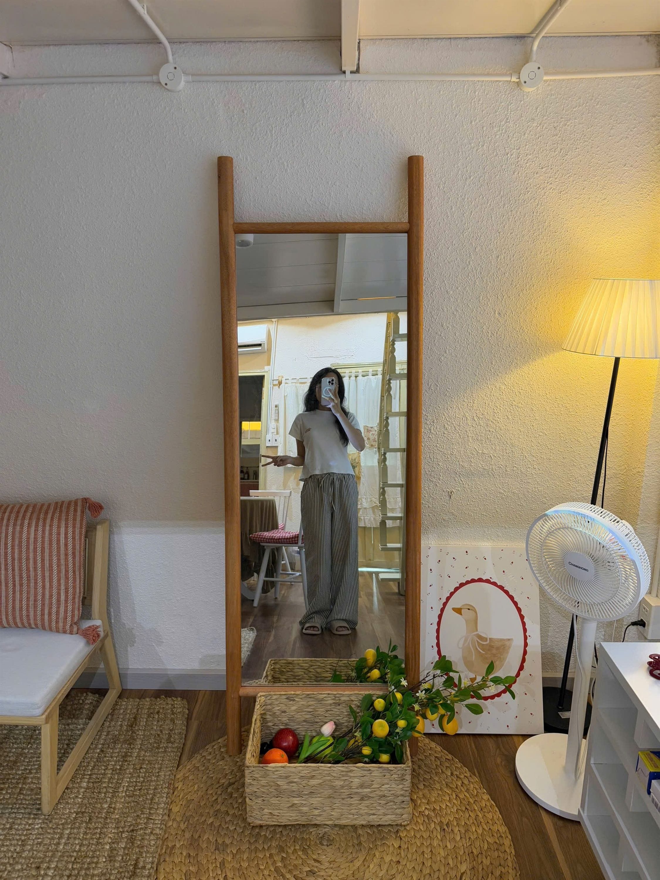 Una habitación con un espejo, un ventilador de pie, una cesta de mimbre con frutas y una lámpara.