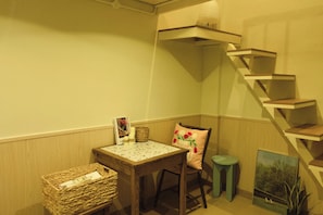 Une pièce d’angle avec une table en bois, une chaise, un petit tabouret, un panier en osier, un tableau encadré et un escalier en bois.