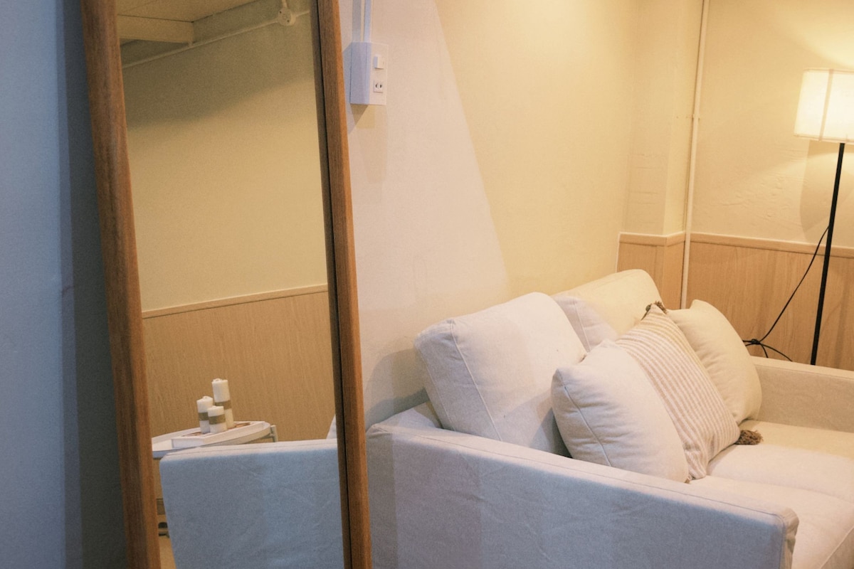Habitación de hotel con un espejo grande, un sofá con almohadas, una lámpara y una pequeña mesa con una botella.