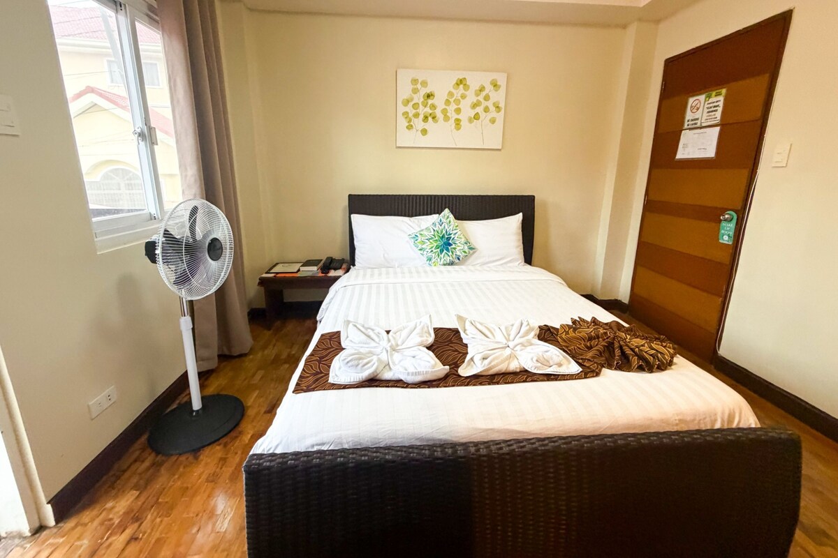 Una habitación de hotel con cama, ventilador de pie, mesita de noche con libros, una obra de arte en la pared y una puerta con un letrero.