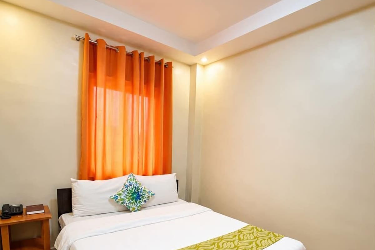 Habitación de hotel con piso de madera, cama con ropa blanca y un plaid verde, mesita de noche con teléfono y libro, y cortinas naranjas.