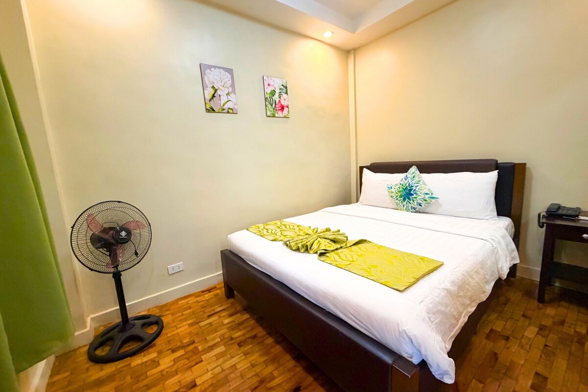 Habitación de hotel con cama, mesita de noche con teléfono, ventilador de pared y dos cuadros con motivos florales.