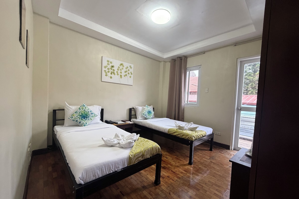 Habitación de hotel con dos camas, suelo de madera, una lámpara en el techo, una ventana con cortinas y una obra de arte colgada en la pared.