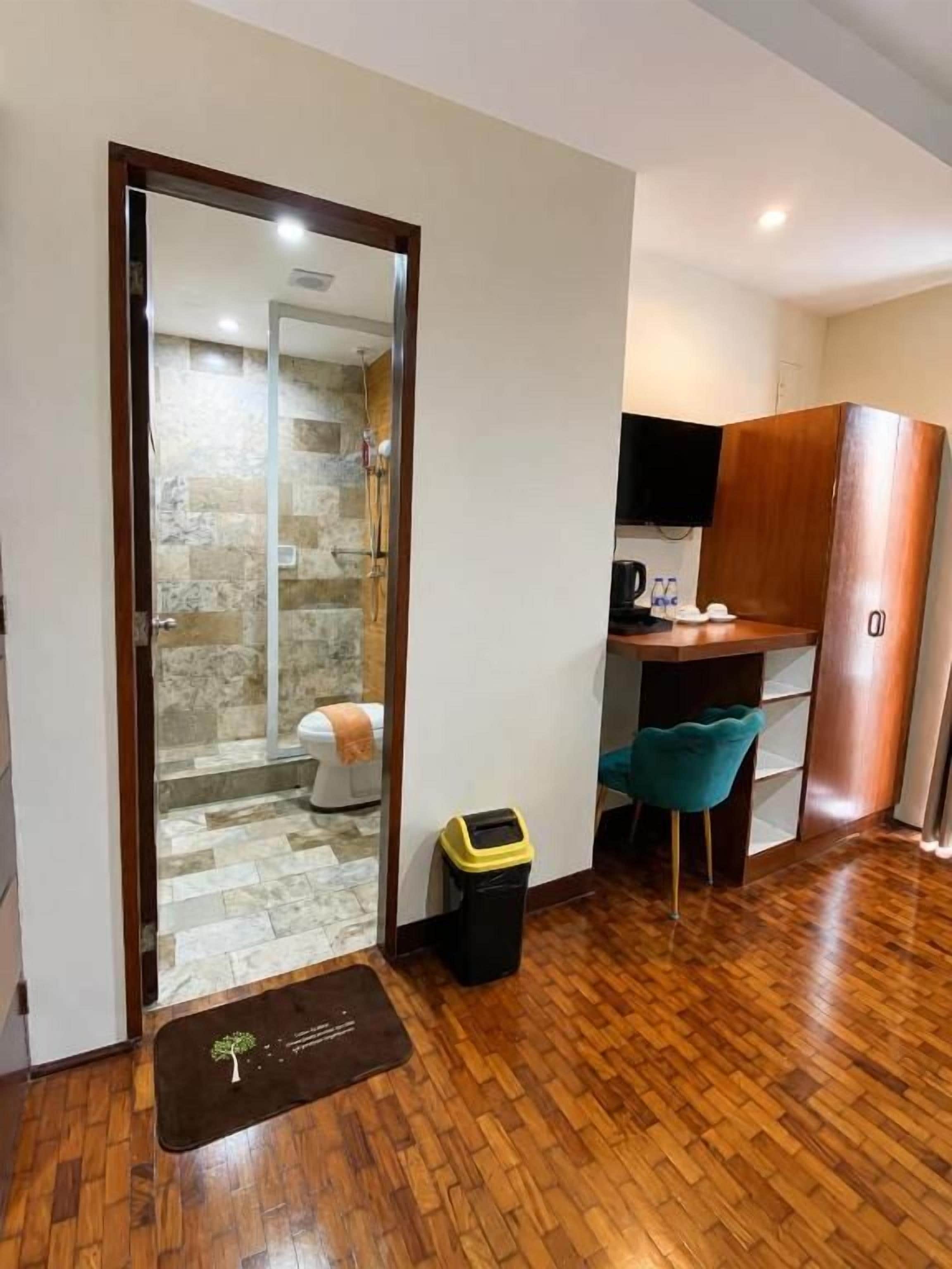 Una habitación compacta con piso de madera, un escritorio pequeño con una silla, un armario y una puerta que da a un baño con ducha.
