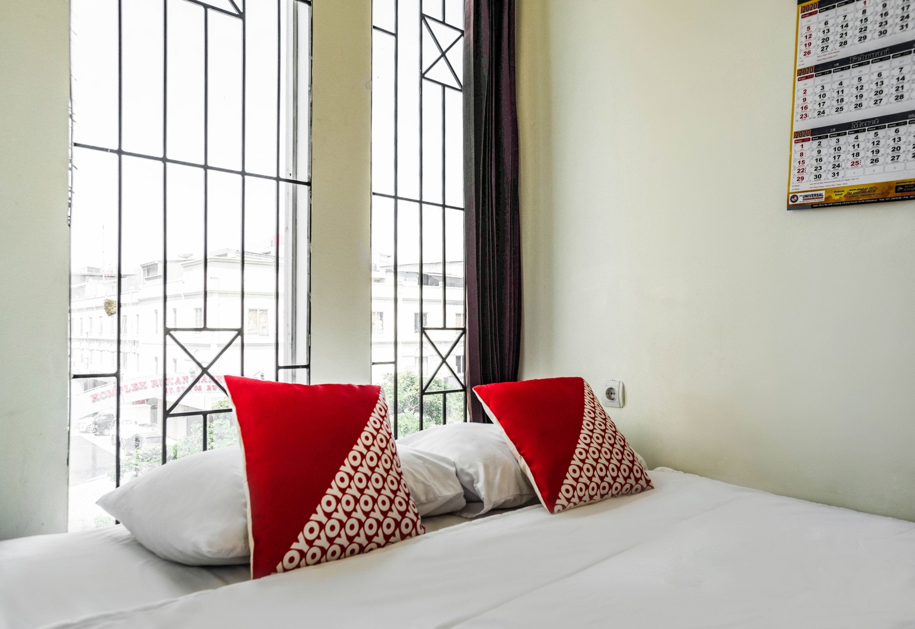 A hotel room with a bed, two pillows, a calendar on the wall, and a window with a view of buildings.