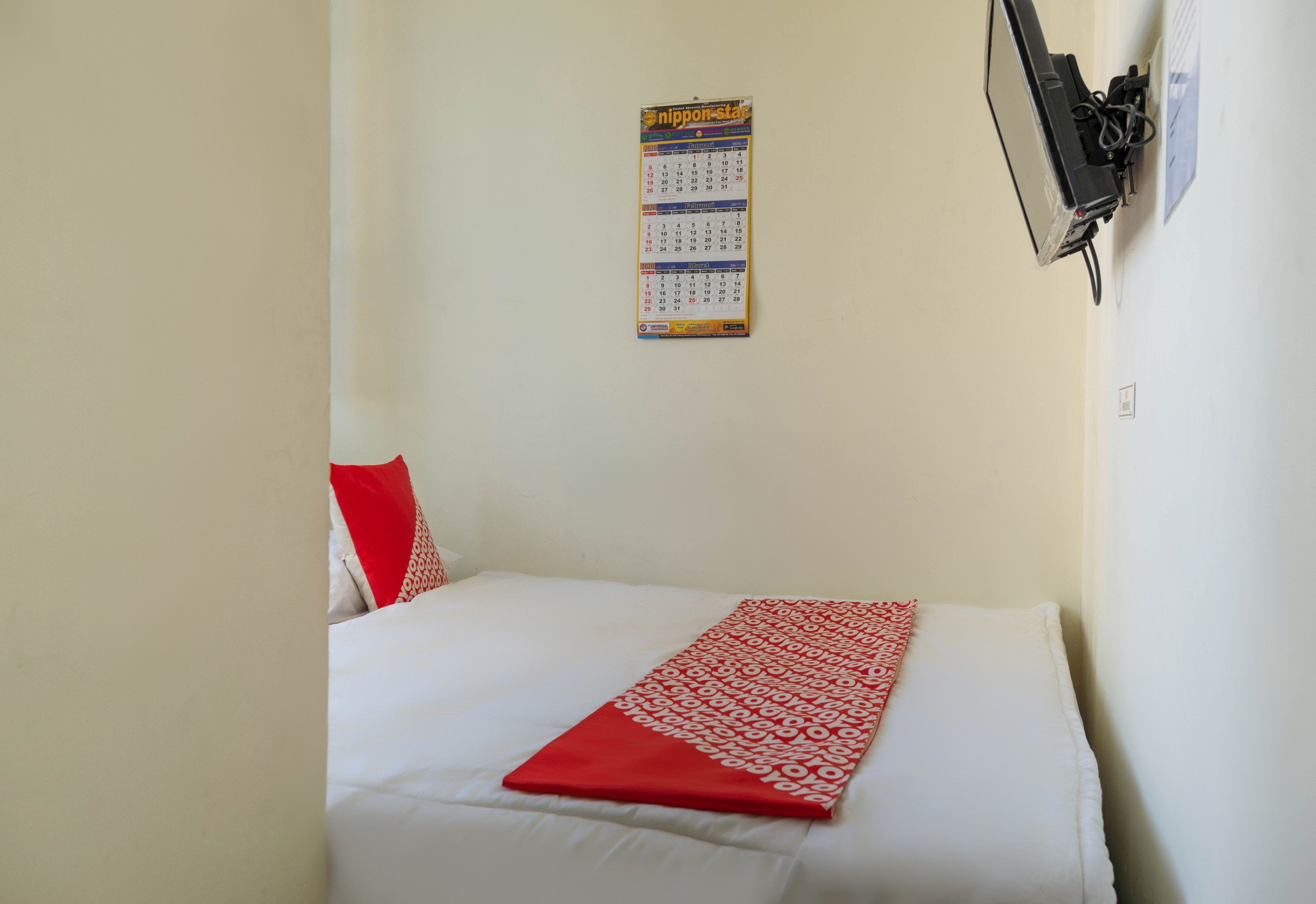 A single bed with a white mattress and a red and white patterned blanket. A calendar is mounted on the wall above the bed.
