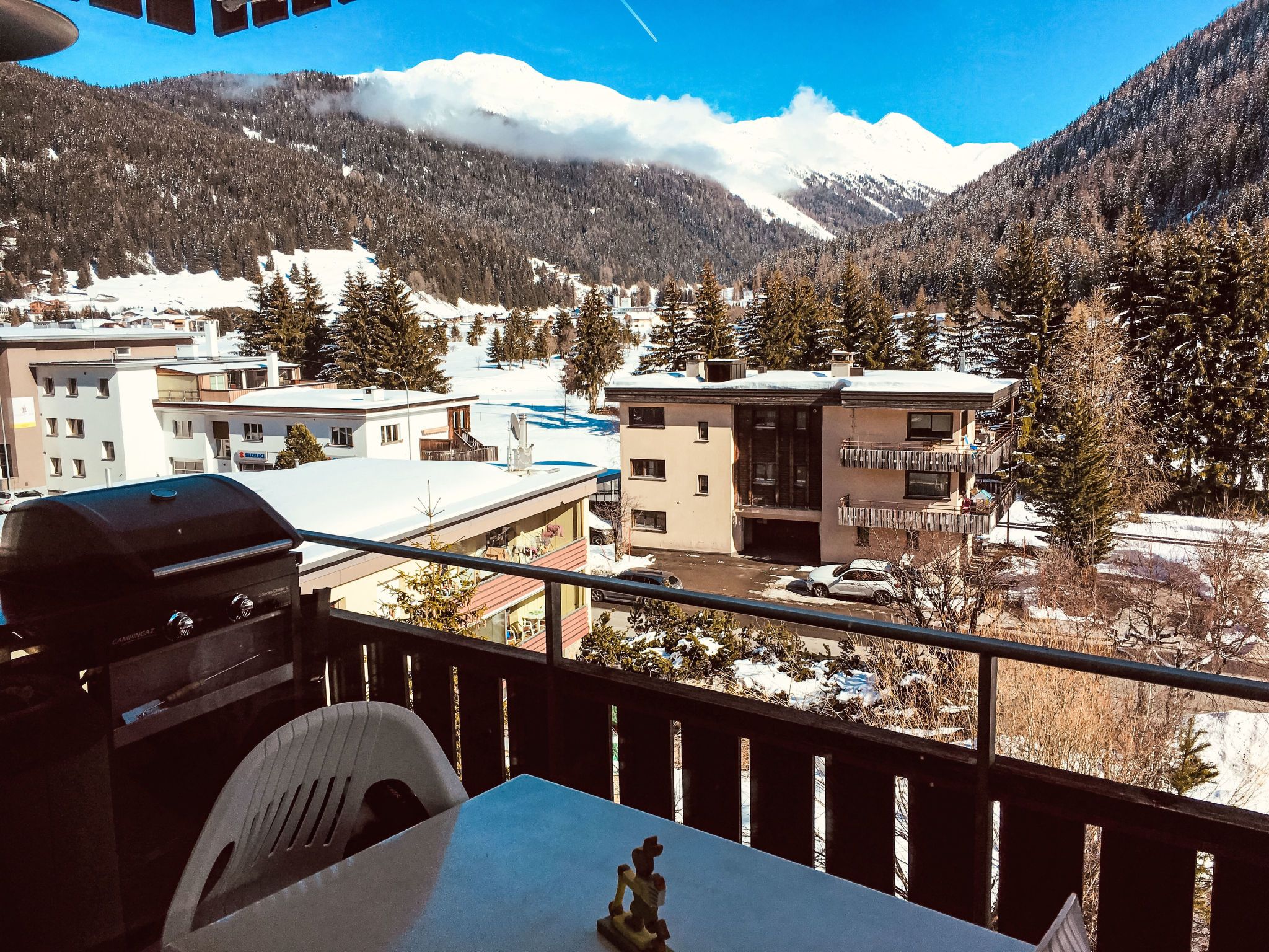 Un balcón con vista a un paisaje montañoso nevado, una parrilla para barbacoa y una mesa con una silla.