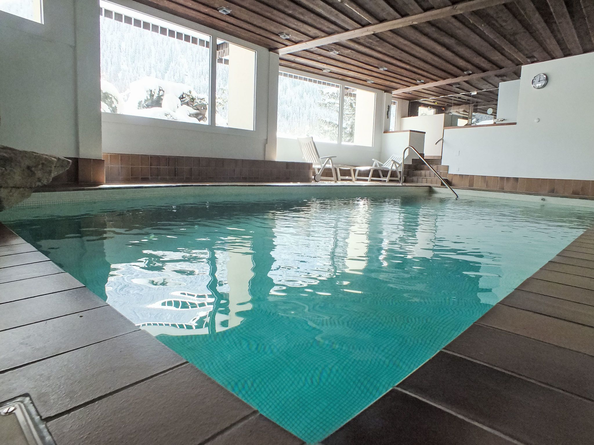 Une piscine intérieure à l’eau claire, dotée d’une terrasse en bois et offrant une vue sur l’extérieur grâce à de grandes fenêtres.
