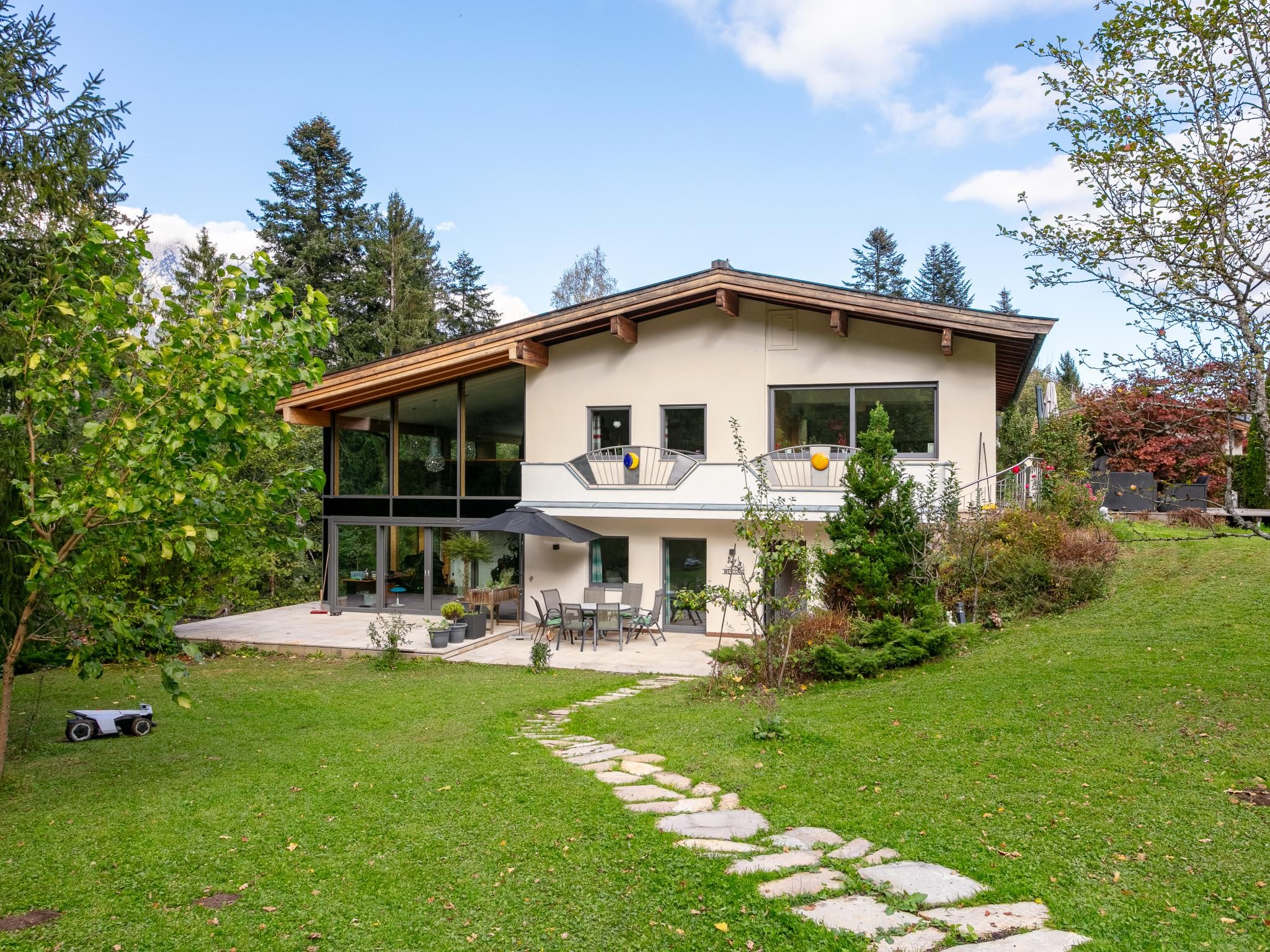 A modern two-story house with a large glass front, a balcony, and a stone pathway leading to the entrance.