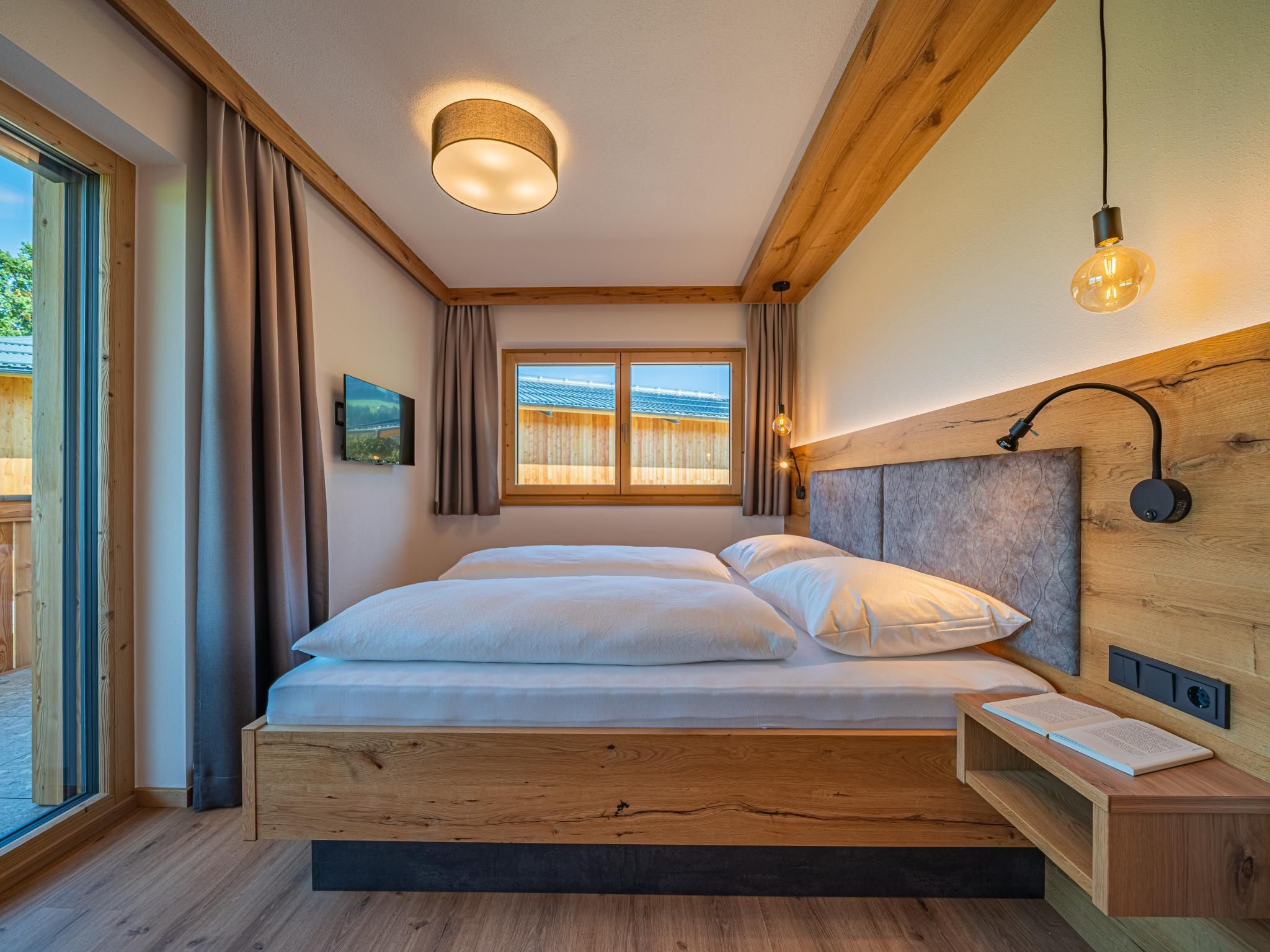 Ein Schlafzimmer mit einem Holzbett, einem Kopfteil mit einer Leuchte, einem Nachttisch mit einem Buch und einem Fenster mit Vorhängen.