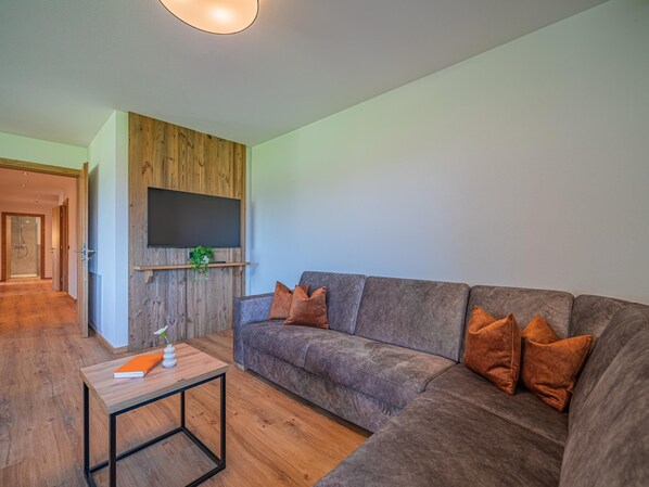 A modern living room with a sectional sofa, a flat-screen TV mounted on a wooden wall, a small table with a book and vase, and a hallway leading to a shower.