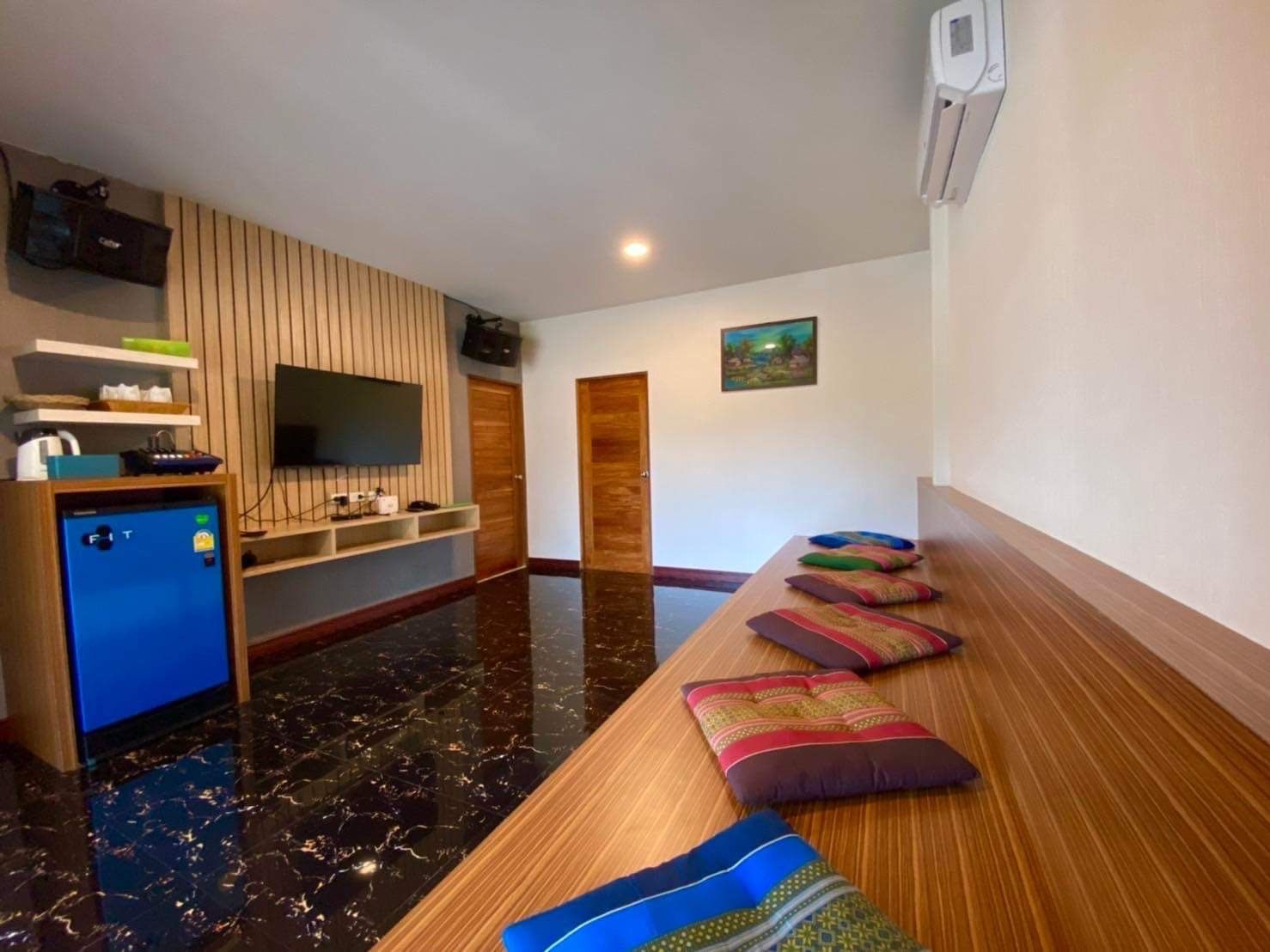 A hotel room with a long wooden bench, a flat-screen TV mounted on the wall, a small refrigerator, and a wooden door.