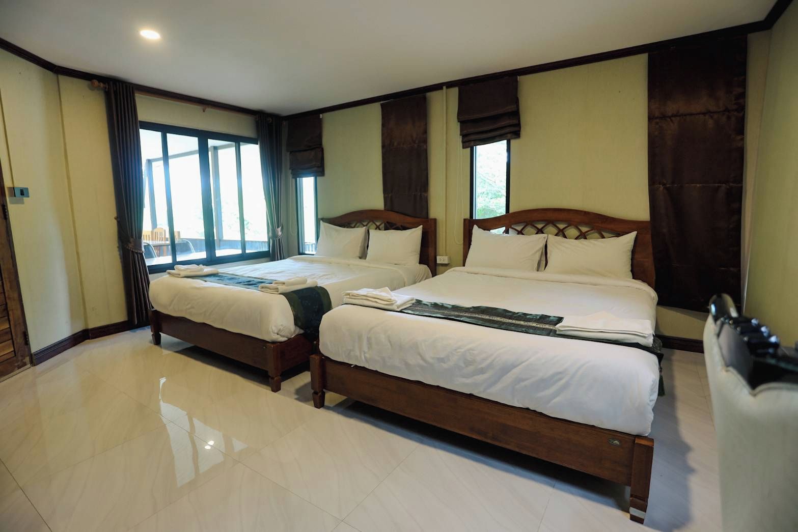 Une chambre d’hôtel avec deux lits en bois, une grande fenêtre avec des rideaux et un sol carrelé.