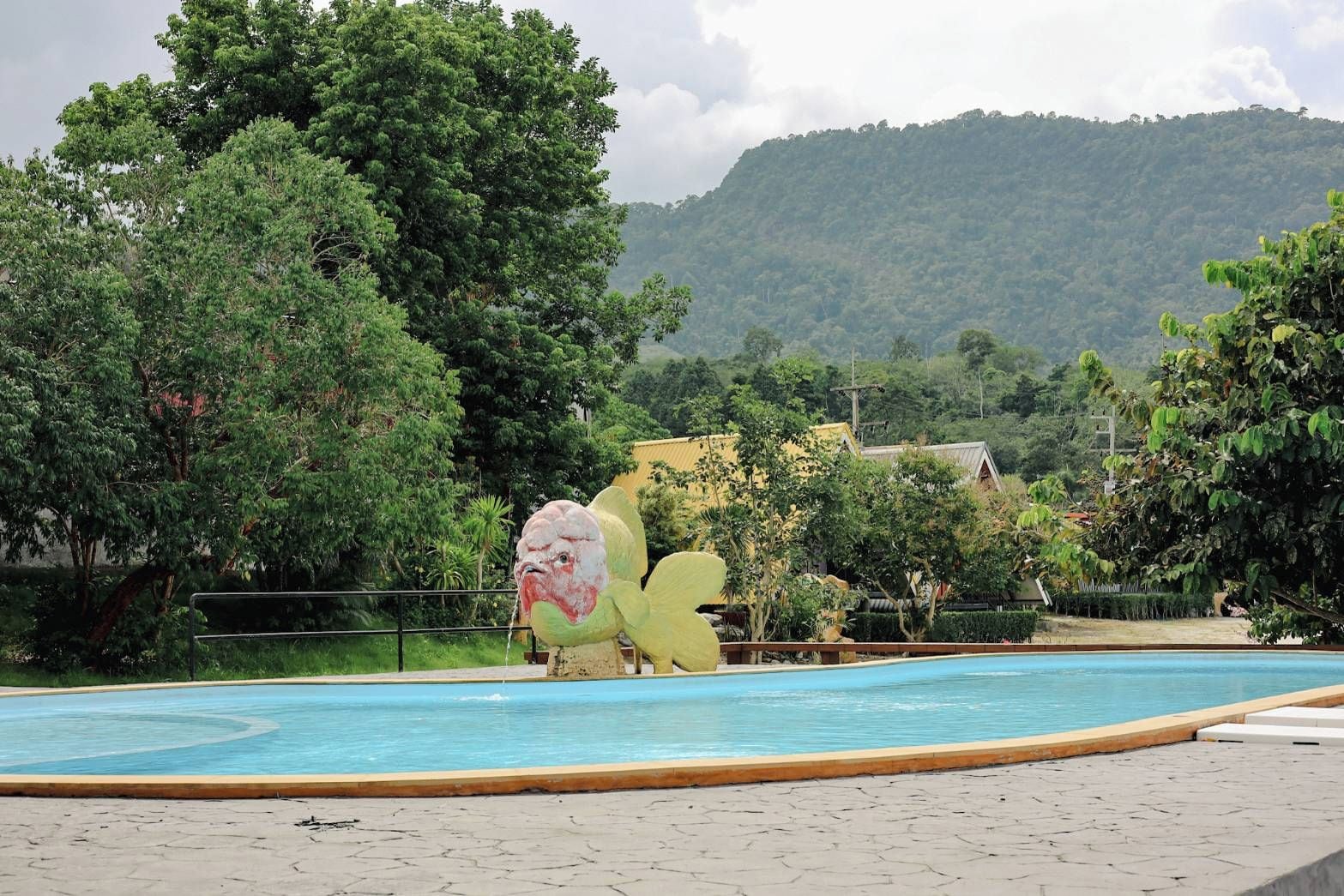 A swimming pool with a large bird sculpture in the center, surrounded by lush greenery and a hilly landscape in the background.