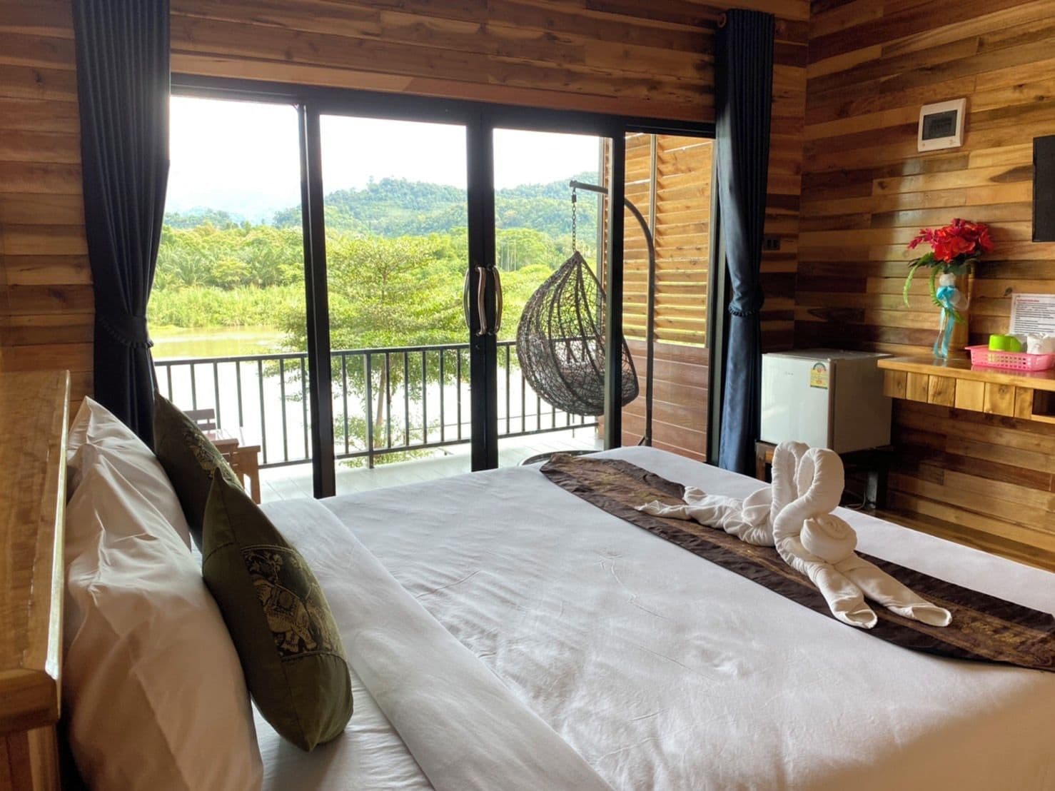 A hotel room with a bed, a hanging chair, a small table, and a view of greenery outside.