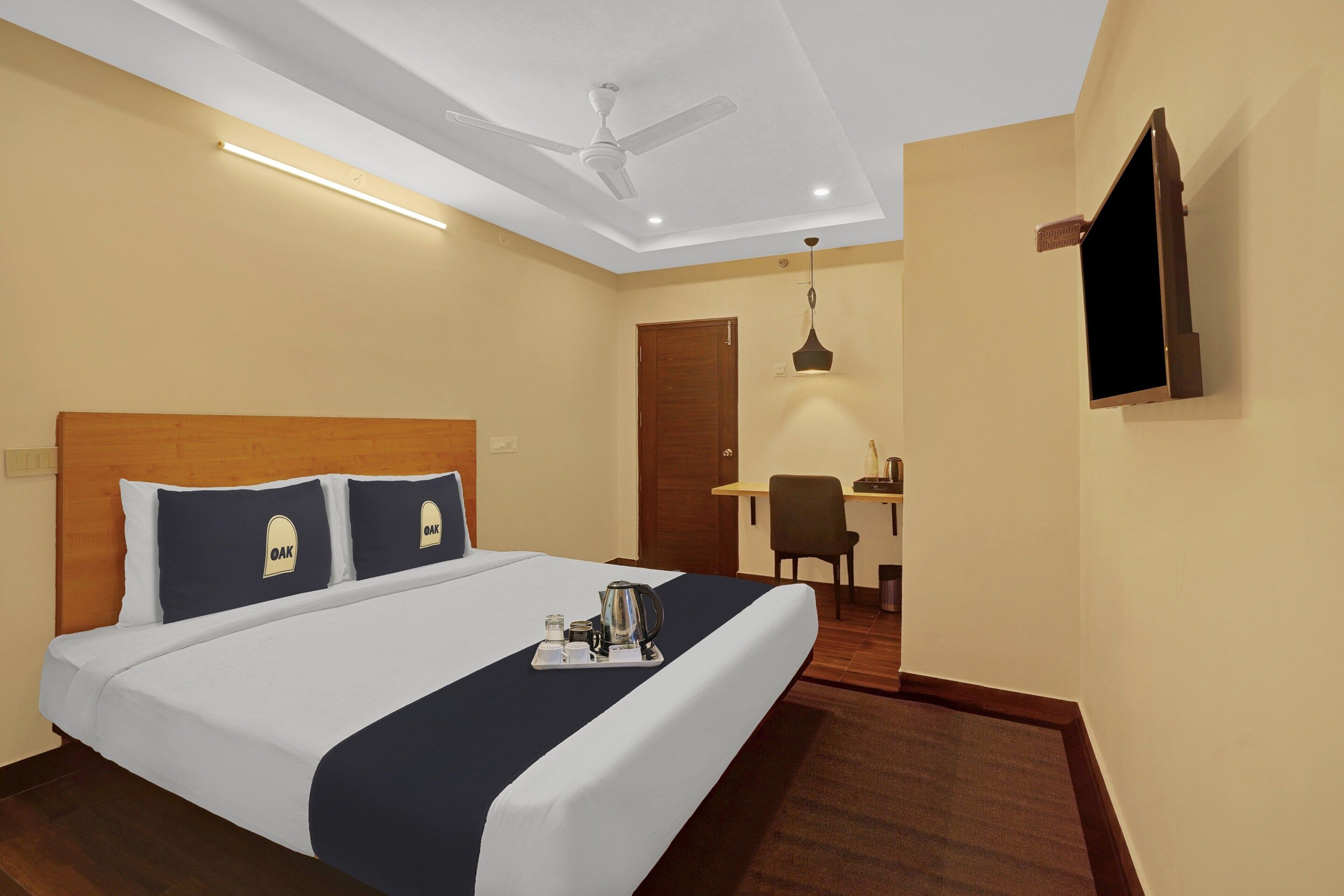 Ein Hotelzimmer mit einem Bett, einem Schreibtisch, einem Sessel, einem Deckenventilator und einem an der Wand befestigten Fernseher.