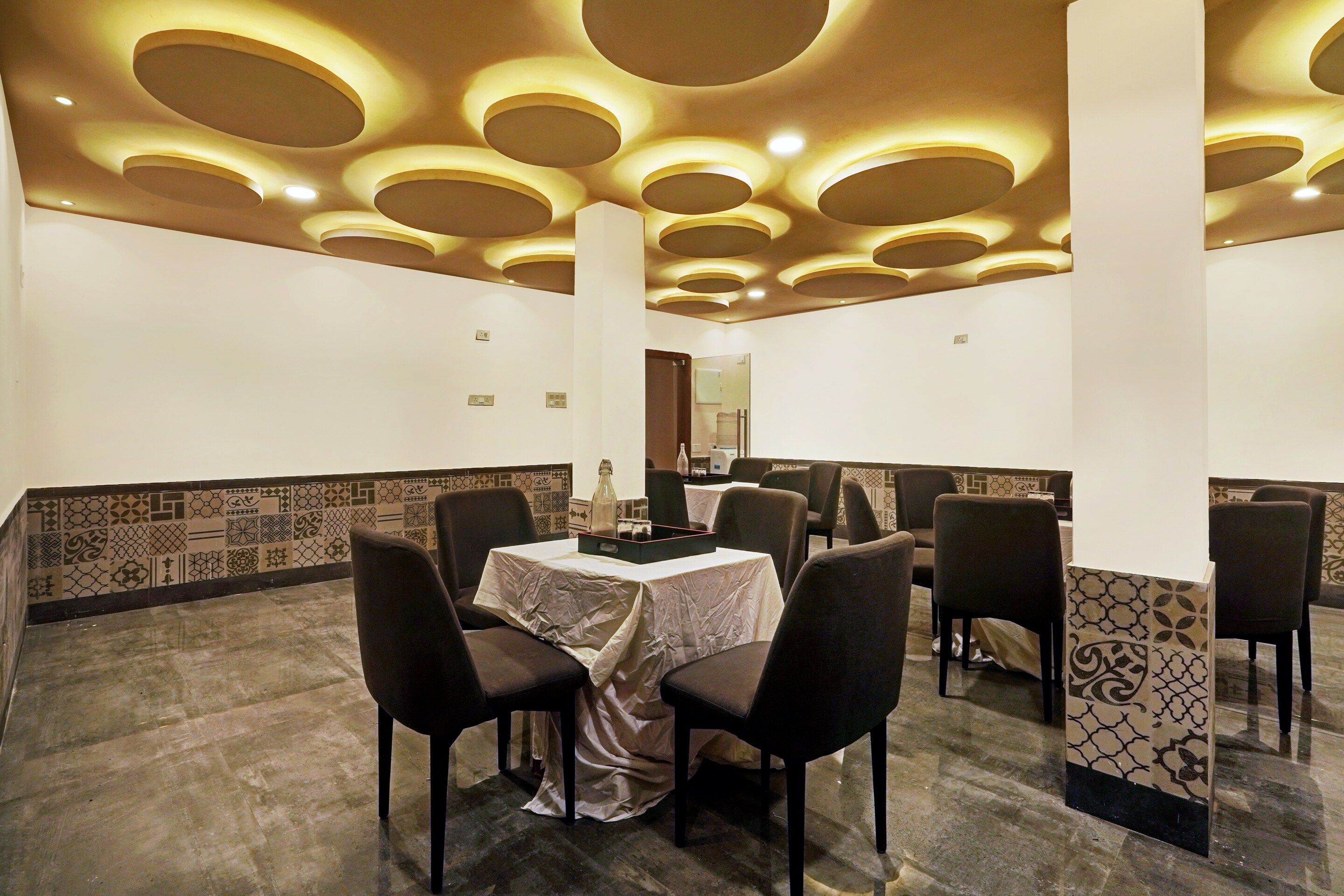 Un espace repas doté d’un plafond haut, agrémenté d’un éclairage encastré circulaire, d’une table centrale entourée de chaises et de carreaux muraux à motifs.