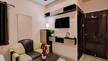 Una sala de estar con sofá, una mesa de centro con un florero, un televisor de pantalla plana montado en la pared, un estante pequeño con una planta y una puerta con pomo.