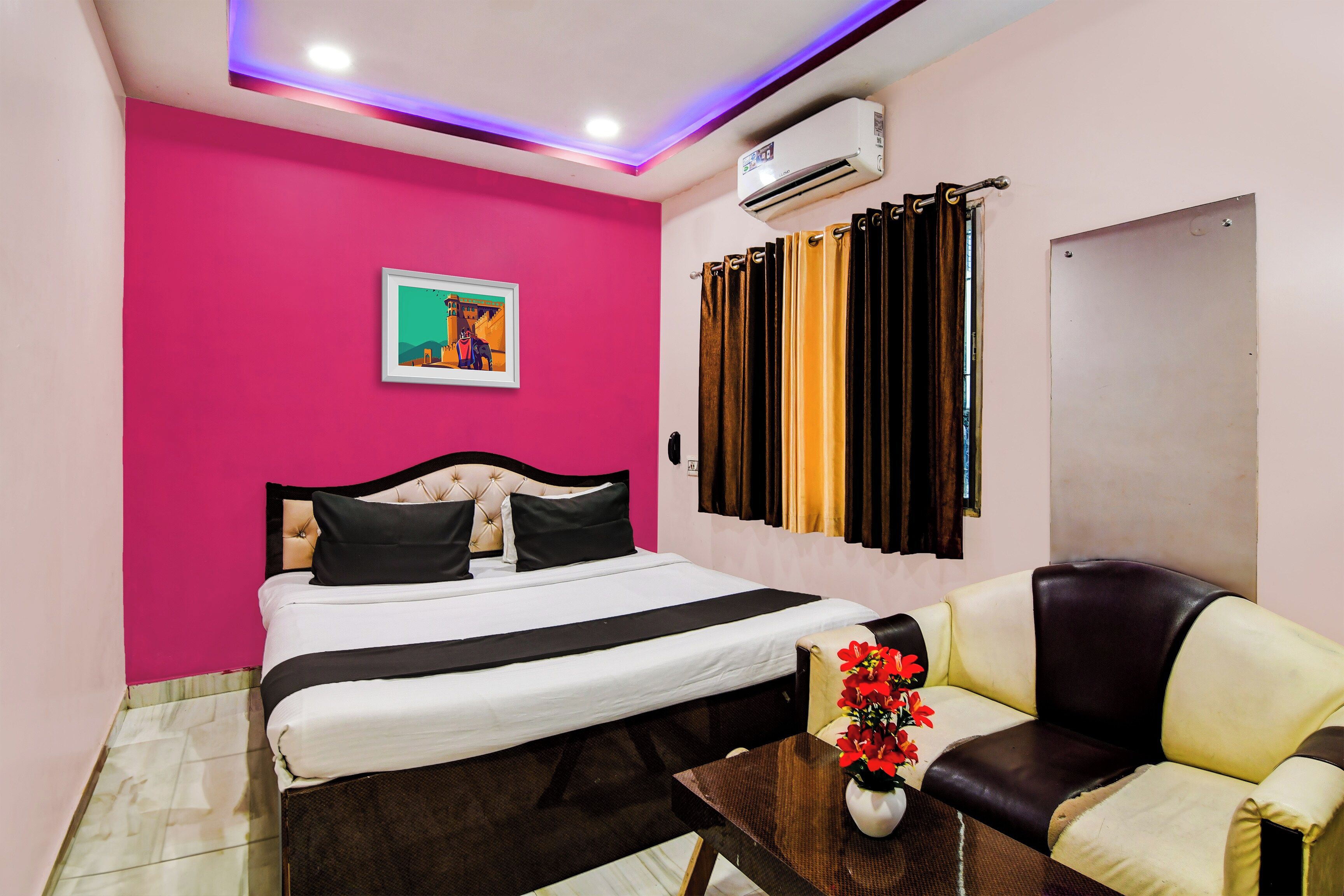 Habitación de hotel con cama, sofá, mesa con un jarrón de flores, un cuadro en la pared y un equipo de aire acondicionado.