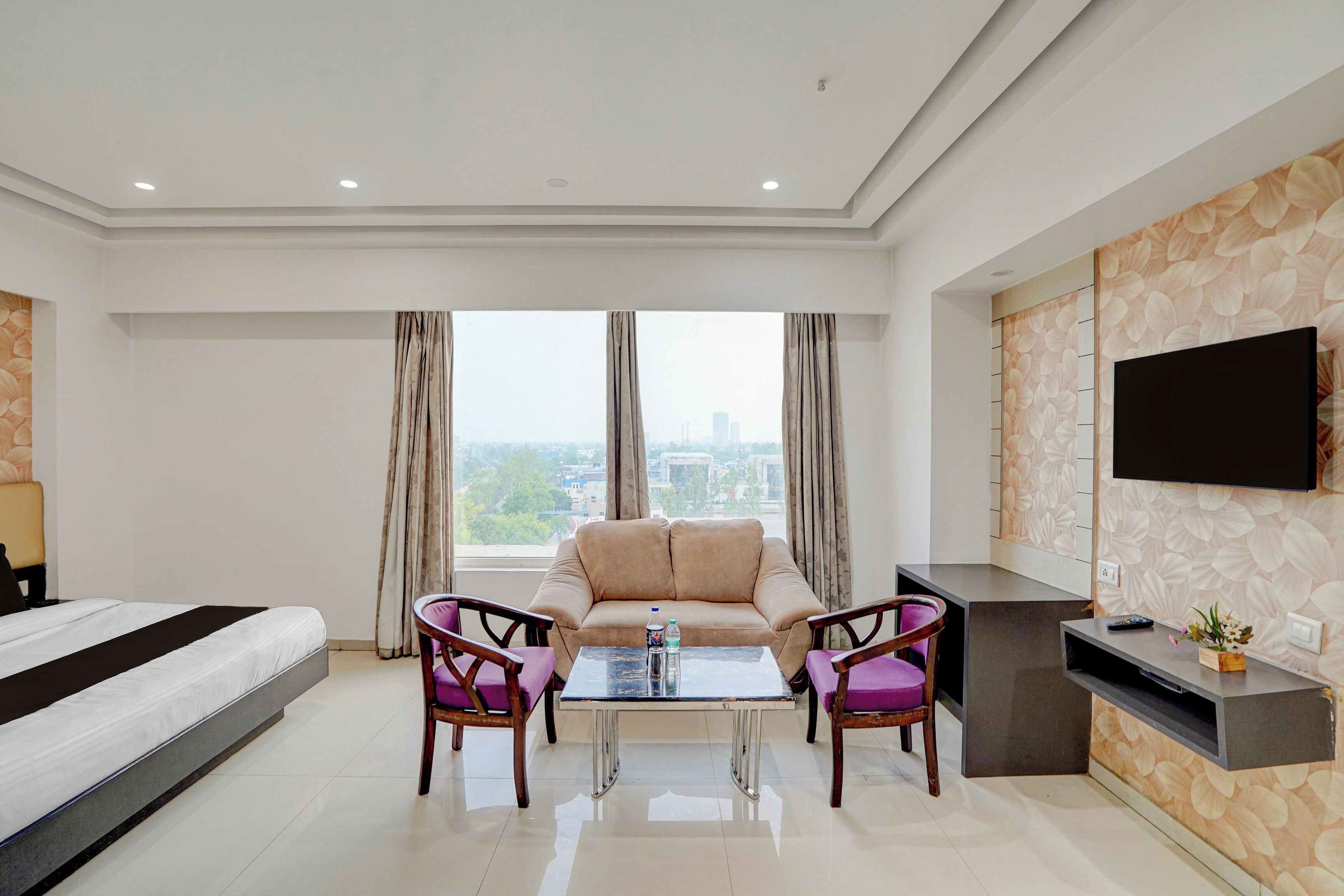 Habitación de hotel con cama, sofá, dos sillones, una mesita de centro, televisor de pantalla plana montado en la pared y un amplio ventanal con cortinas.