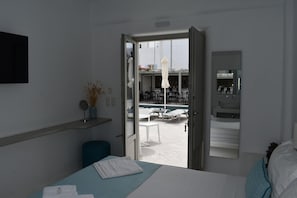 A hotel room with a bed, a bathroom with a sink and mirror, and an open door leading to a pool area with lounge chairs. - Angels Pillow (Paros)