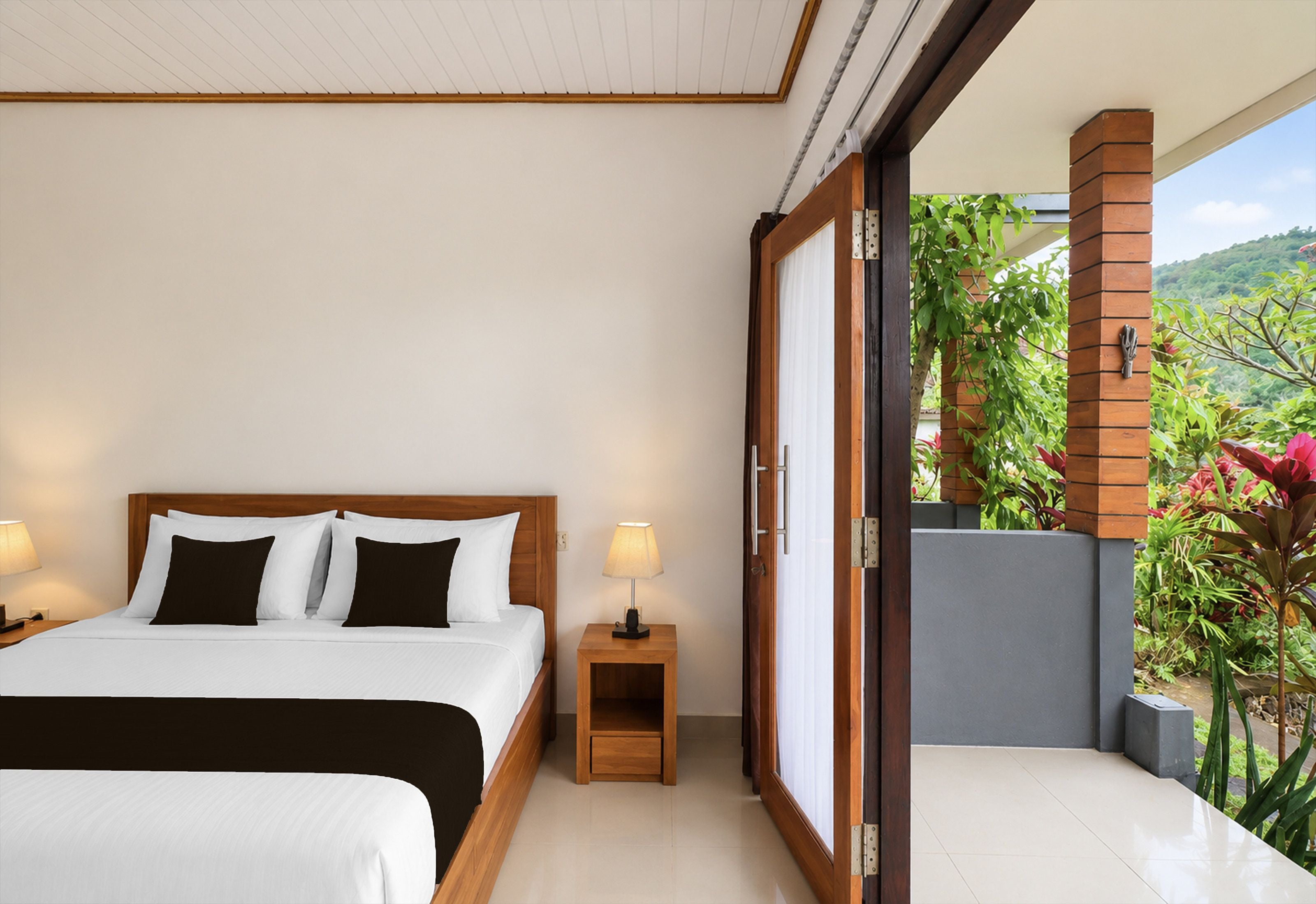 Habitación de hotel con una cama grande, una mesita de noche, una lámpara y un ventanal corredero que da a un balcón con plantas.