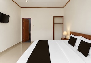 Una habitación de hotel con cama, televisor montado en la pared, puerta y mesita de noche con lámpara.