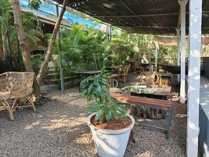 Un área de estar al aire libre con mesas y sillas, rodeada de follaje y una estructura cubierta.