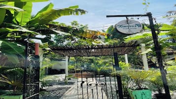 Une entrée fermée par un portail, avec une enseigne indiquant « Xavier House, bar-restaurant et maison d’hôtes », et un jardin aux allures tropicales.