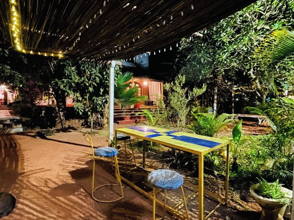 Un área de estar al aire libre con una mesa y sillas, rodeada de abundante vegetación y una estructura cubierta.