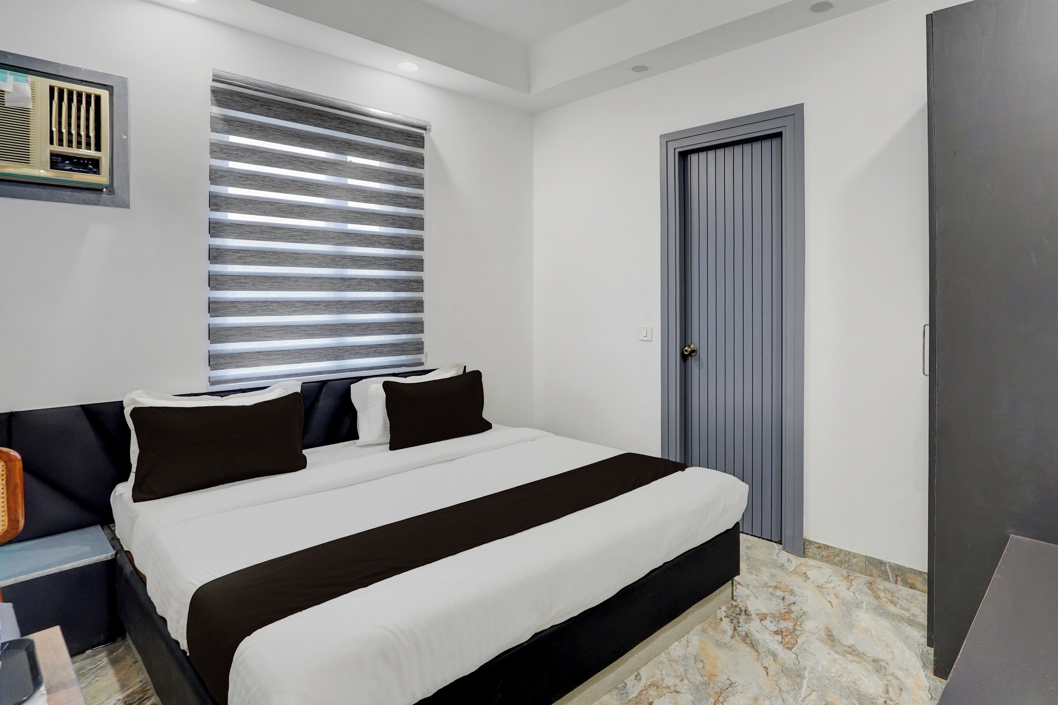 Un dormitorio con cama, mesita de noche, ventana con persianas, puerta y aire acondicionado instalado en la pared.