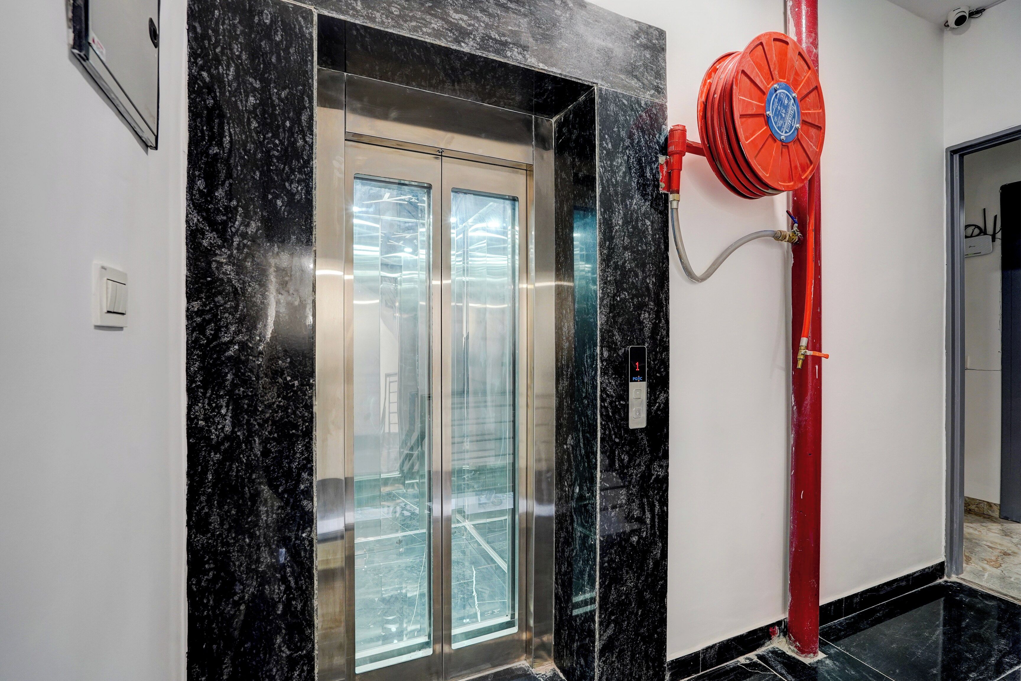 An elevator with a glass door, a red fire hose reel, and a visible electrical outlet on the wall.