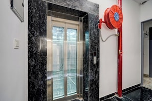 Un ascensor con puerta de vidrio, un carrete rojo para manguera de incendios y un enchufe eléctrico visible en la pared.
