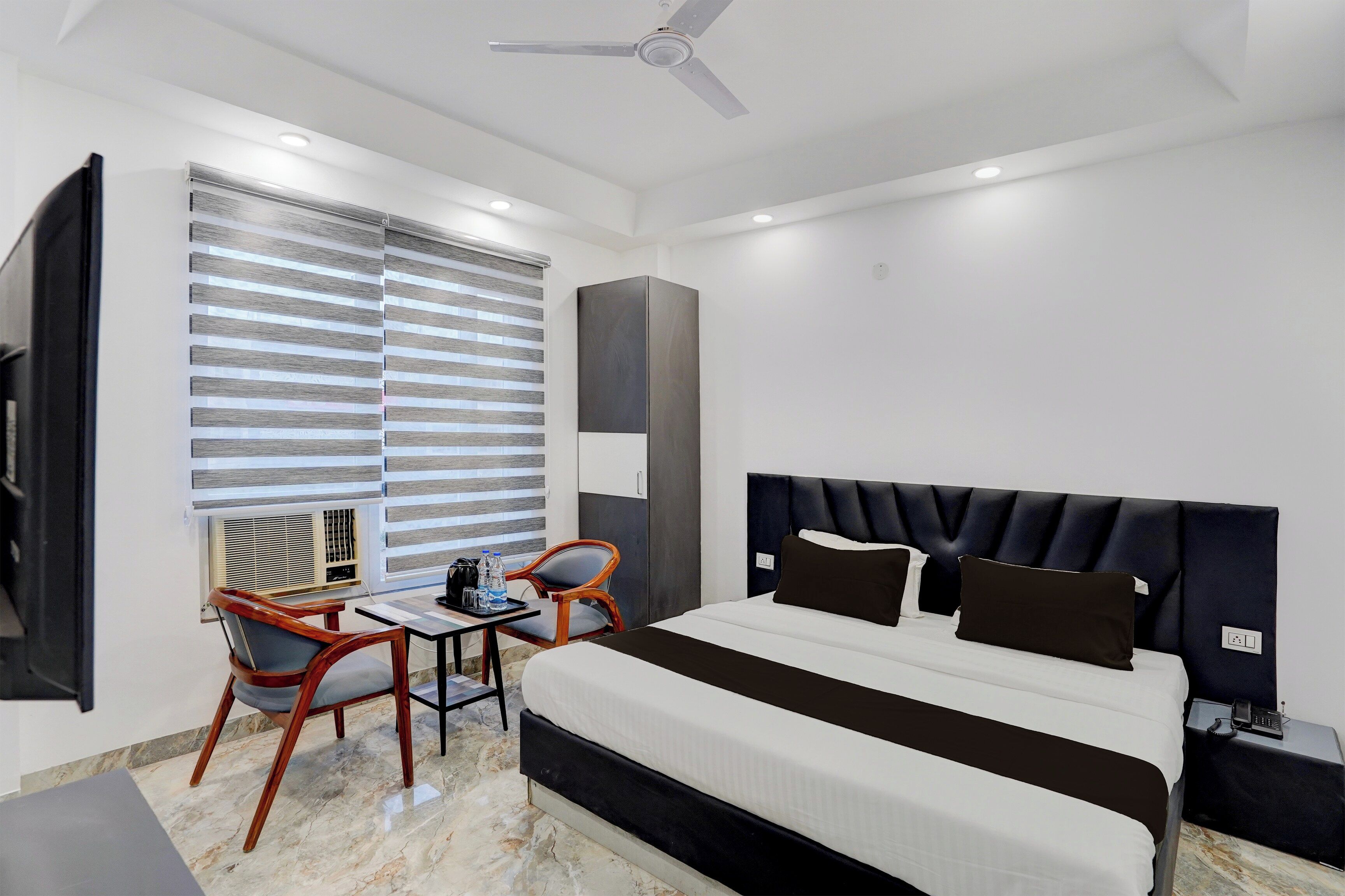 Une chambre d’hôtel moderne équipée d’un lit, d’un bureau, de deux chaises, d’un ventilateur de plafond et d’une grande fenêtre avec des stores.