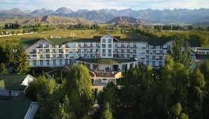 Un hotel grande de varios pisos con fachada simétrica, patio central y vista a montañas al fondo.