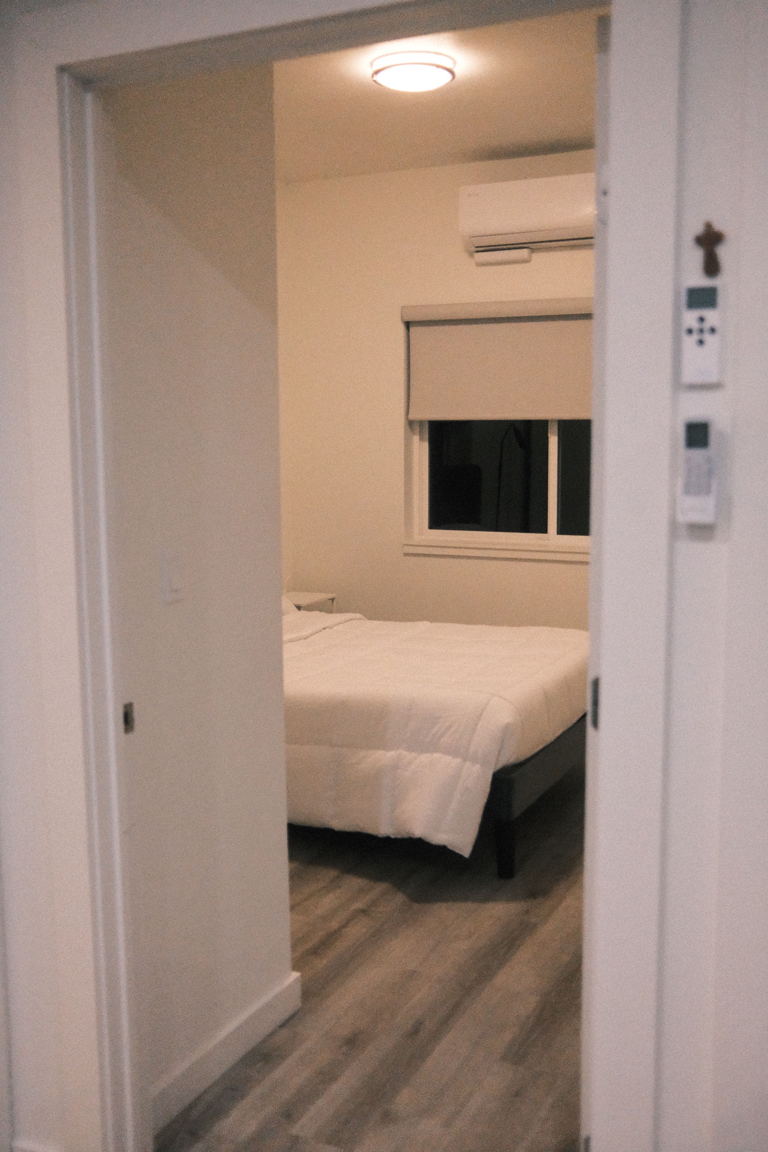 A narrow hallway leading to a bedroom with a single bed, a window, and an air conditioning unit.