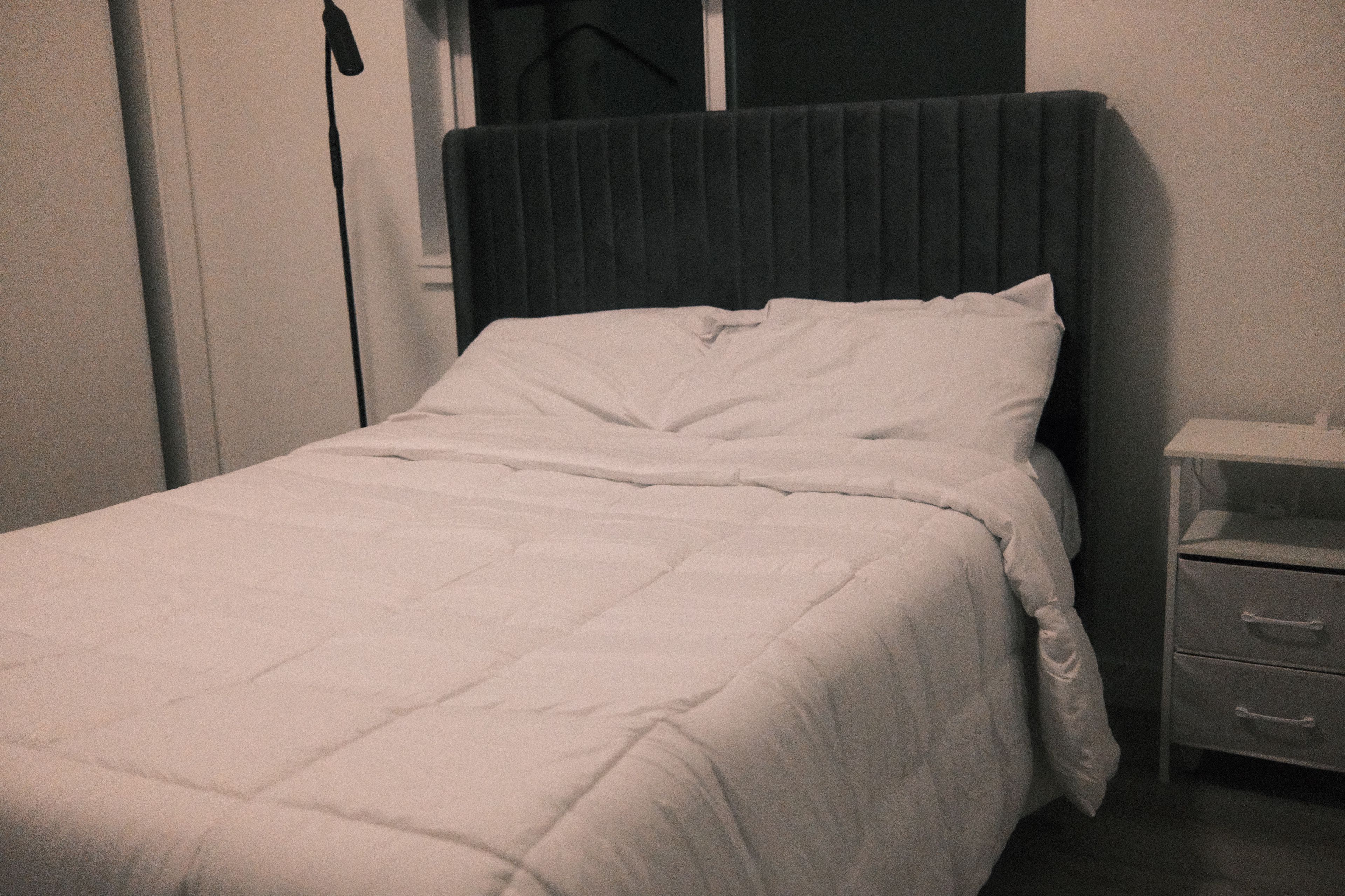 Una cama individual con cabecera oscura en una habitación con mesita de noche y cajón.